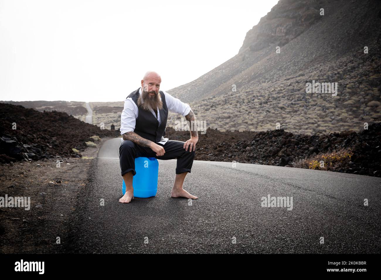 Man with suit is sitting completely desperate and knocked out on a blue gasoline canister somewhere in the nowhere in the volcanic desert at the end o Stock Photo