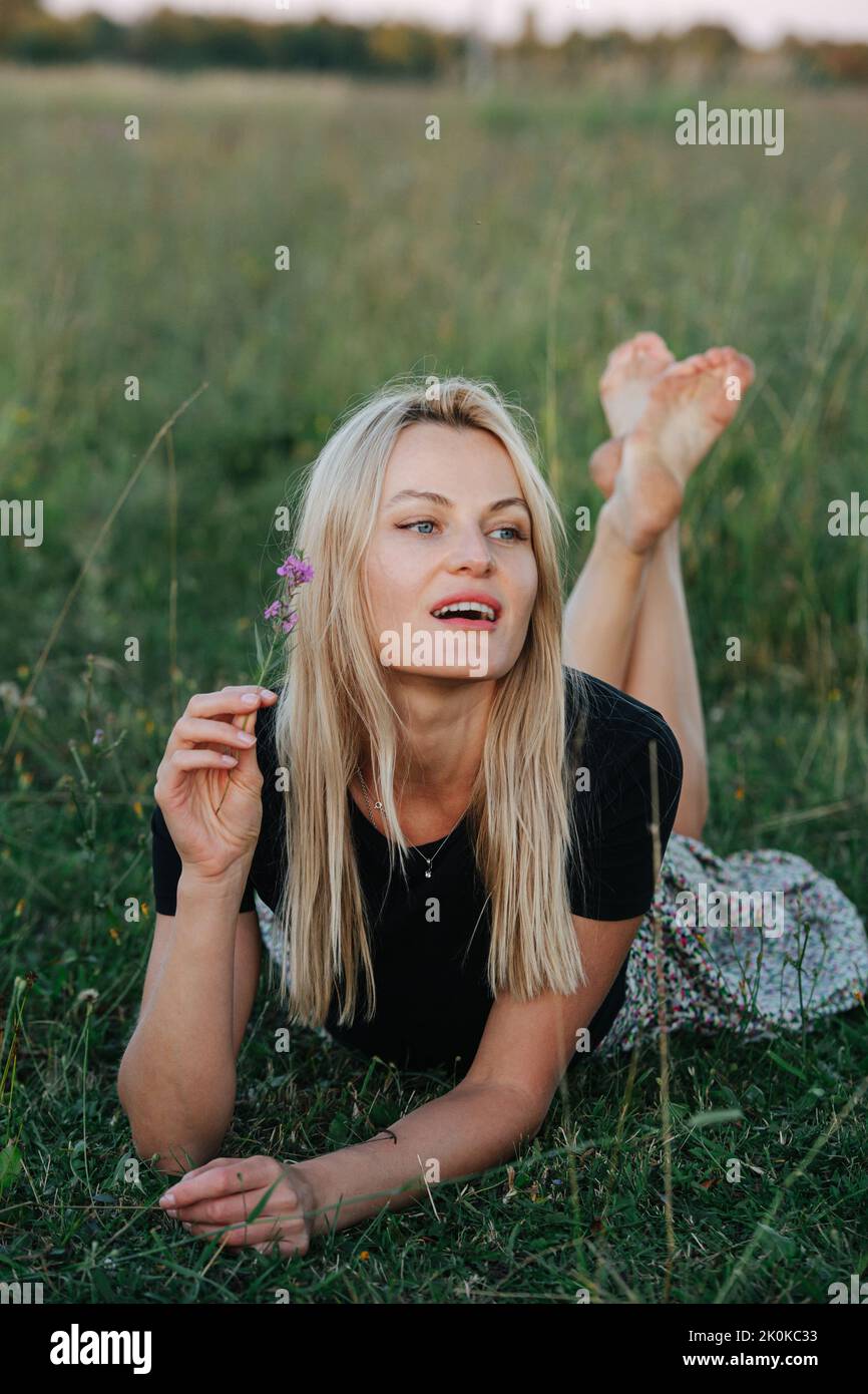 Distracted young blond woman lying on a grass in a field, holding a wild flower Stock Photo