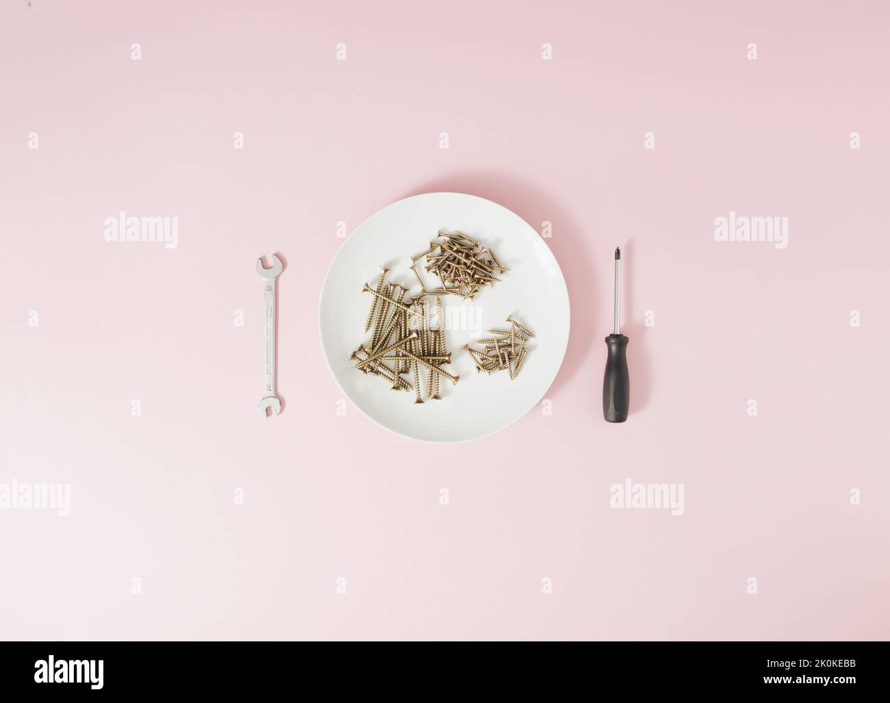 Screws served on a white plate with a screwdriver and a wrench on a pale pink background. Stock Photo