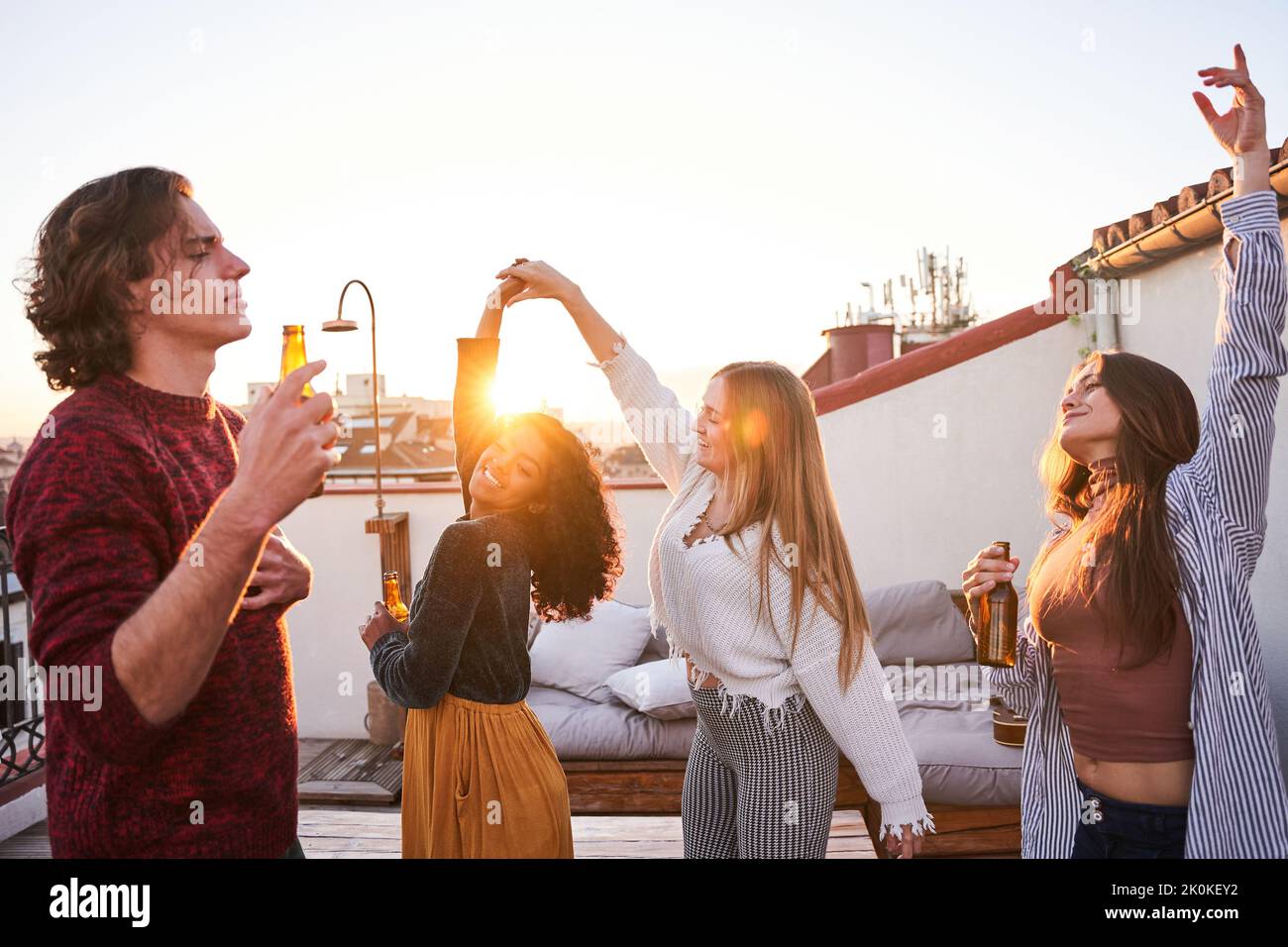Group of happy young multiracial friends in casual clothes drinking cold beer and dancing together while having party on building rooftop at sunset Stock Photo