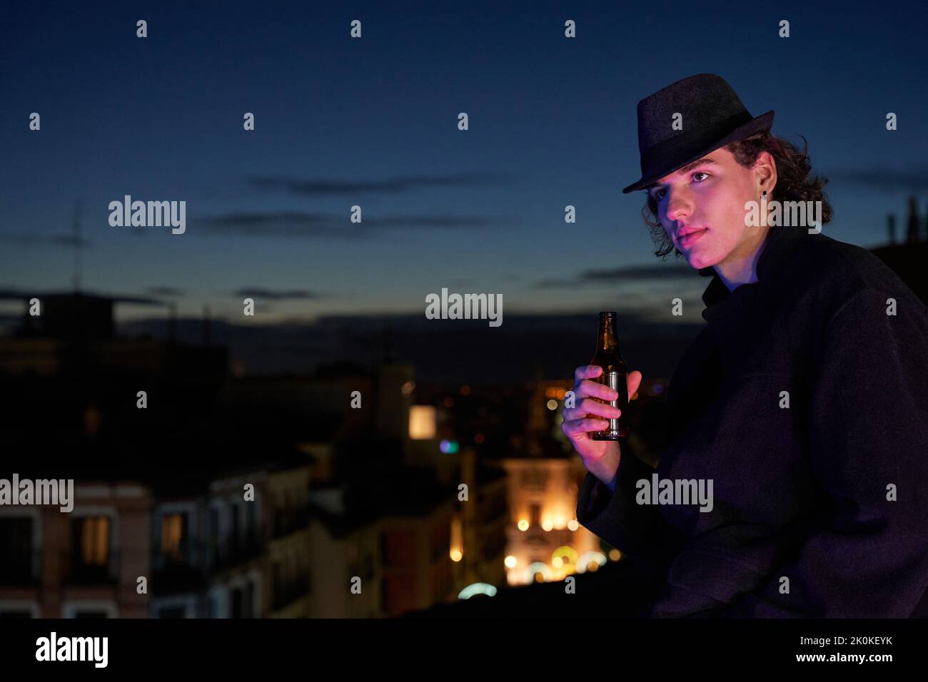 Positive young male in black coat and hat enjoying cold beer and looking away while sitting on rooftop at dusk Stock Photo