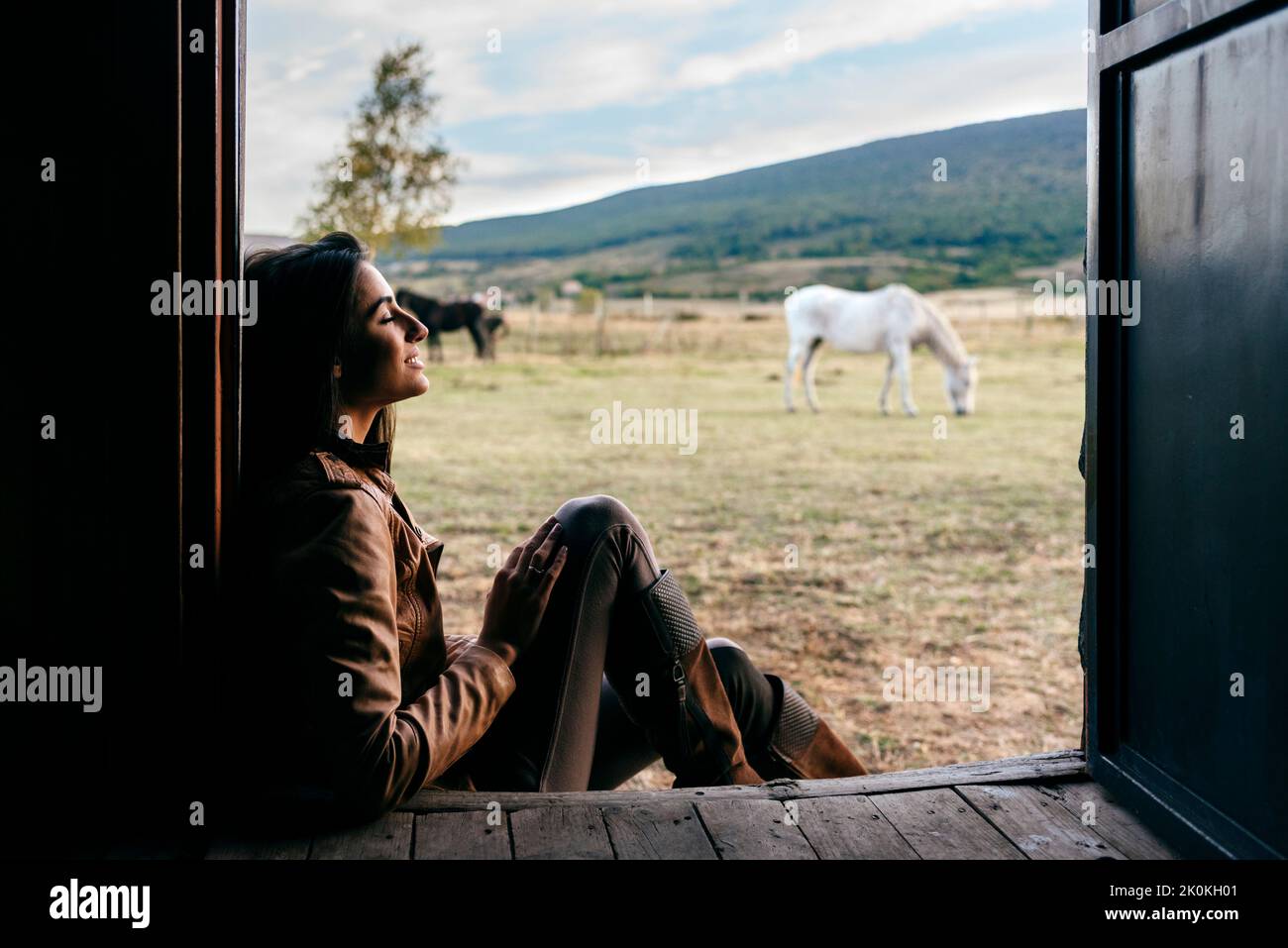 Stylish woman in boots and scarf sitting on porch of old wooden house ...