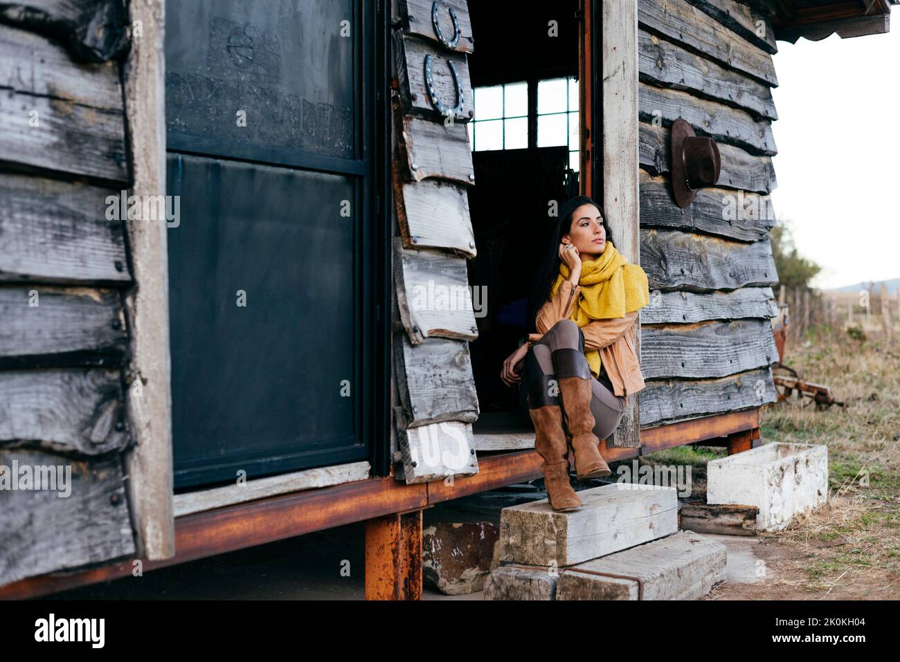 Stylish woman in boots and scarf sitting on porch of old wooden house ...