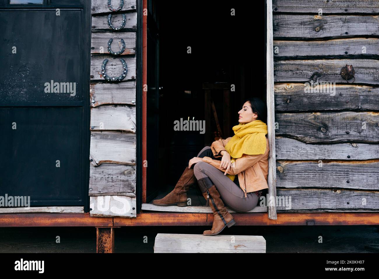 Stylish woman in boots and scarf sitting on porch of old wooden house ...
