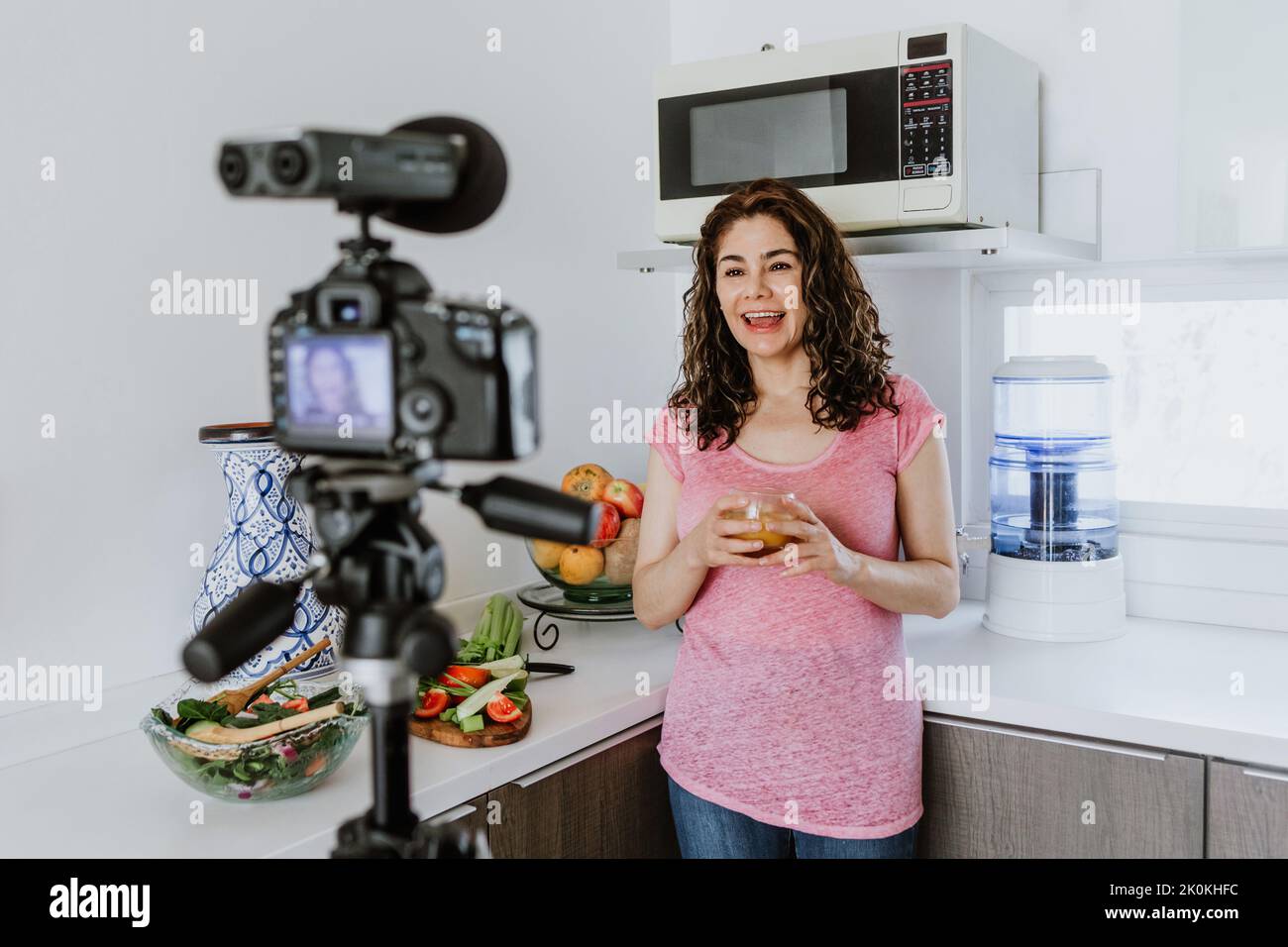 Positive female on casual clothes standing in kitchen and looking at camera on tripod while recording video for vlog about healthy food Stock Photo
