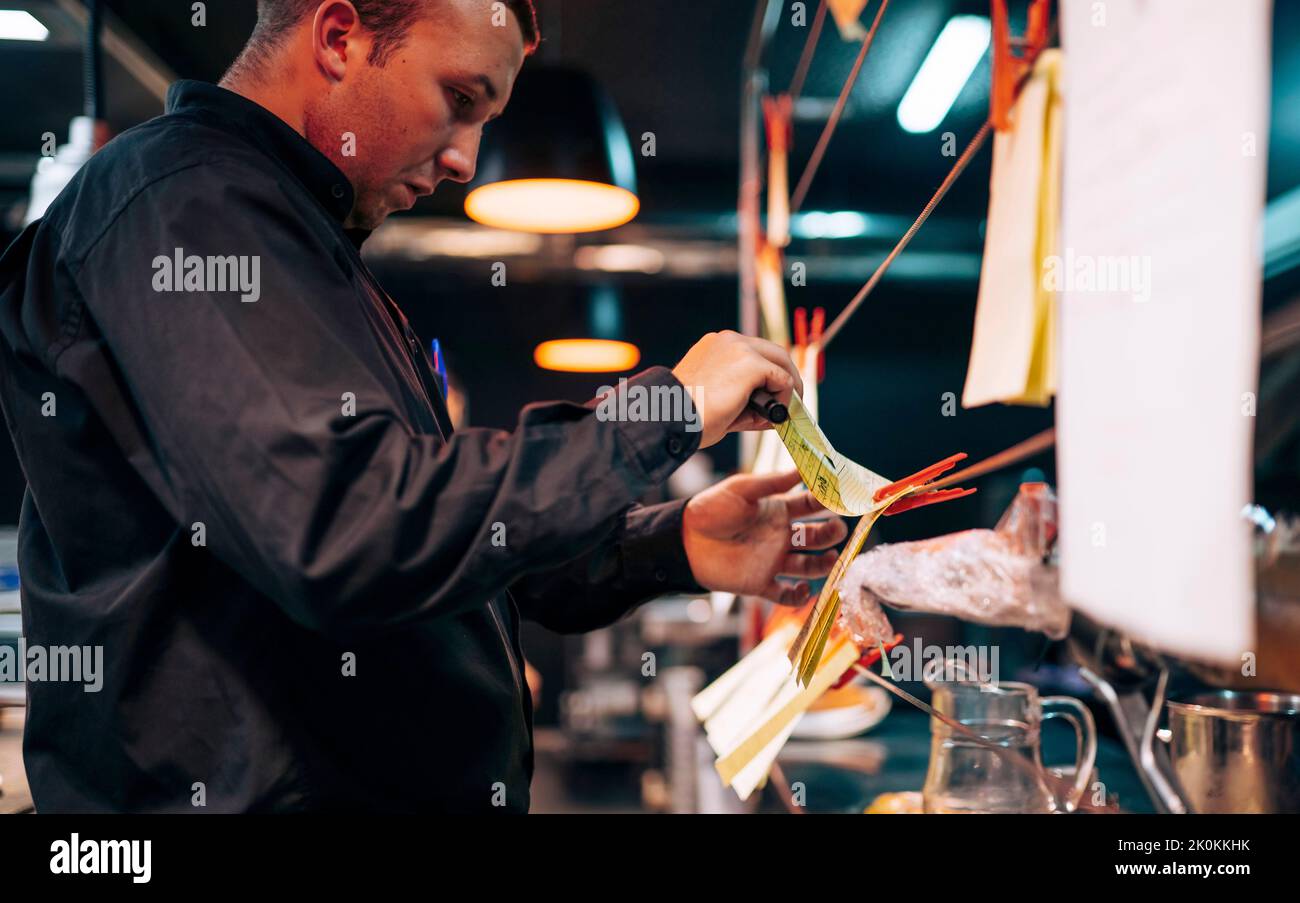 Side view of man in black shirt arranging paper with food orders on strings in kitchen Stock Photo