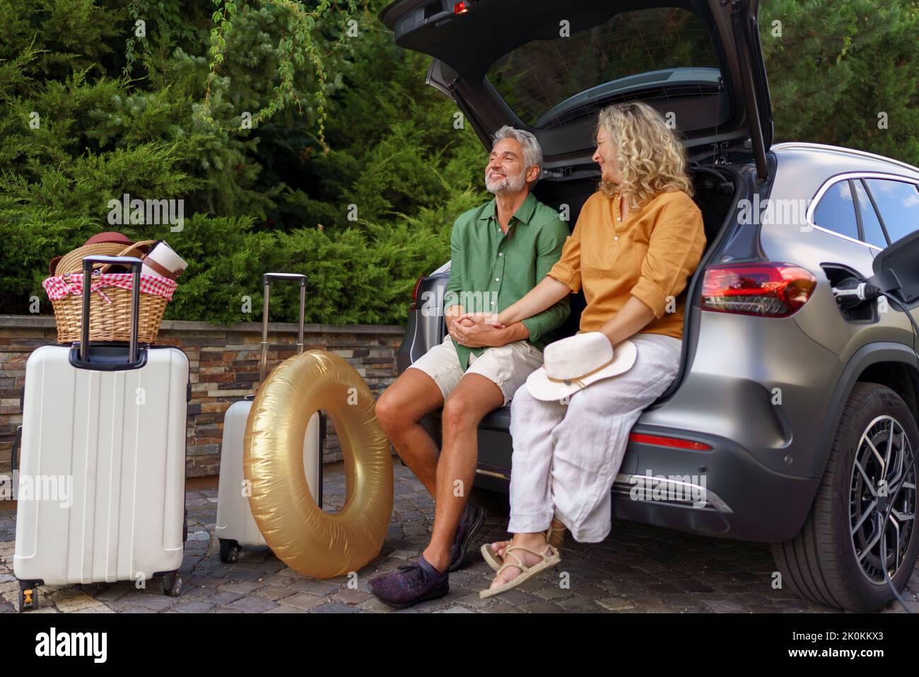 Middle-aged couple sitting in trunk while waiting for charging car before travelling on summer holiday. Stock Photo