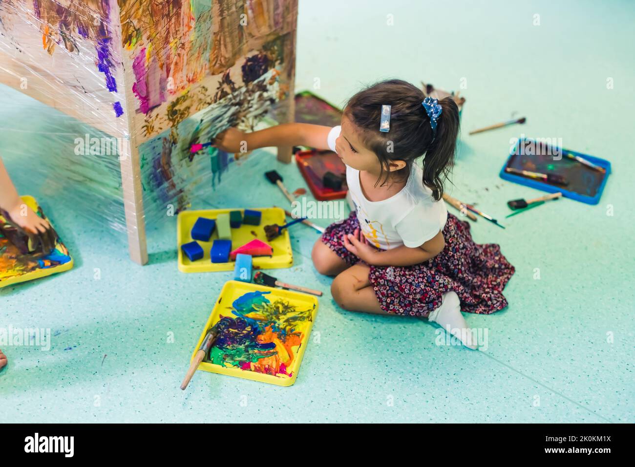Cling film painting. Little girl toddler painting with a sponge and paints on a cling film wrapped all the way round the wooden shelf unit. Creative activity for kids development at the nursery school. High quality photo Stock Photo