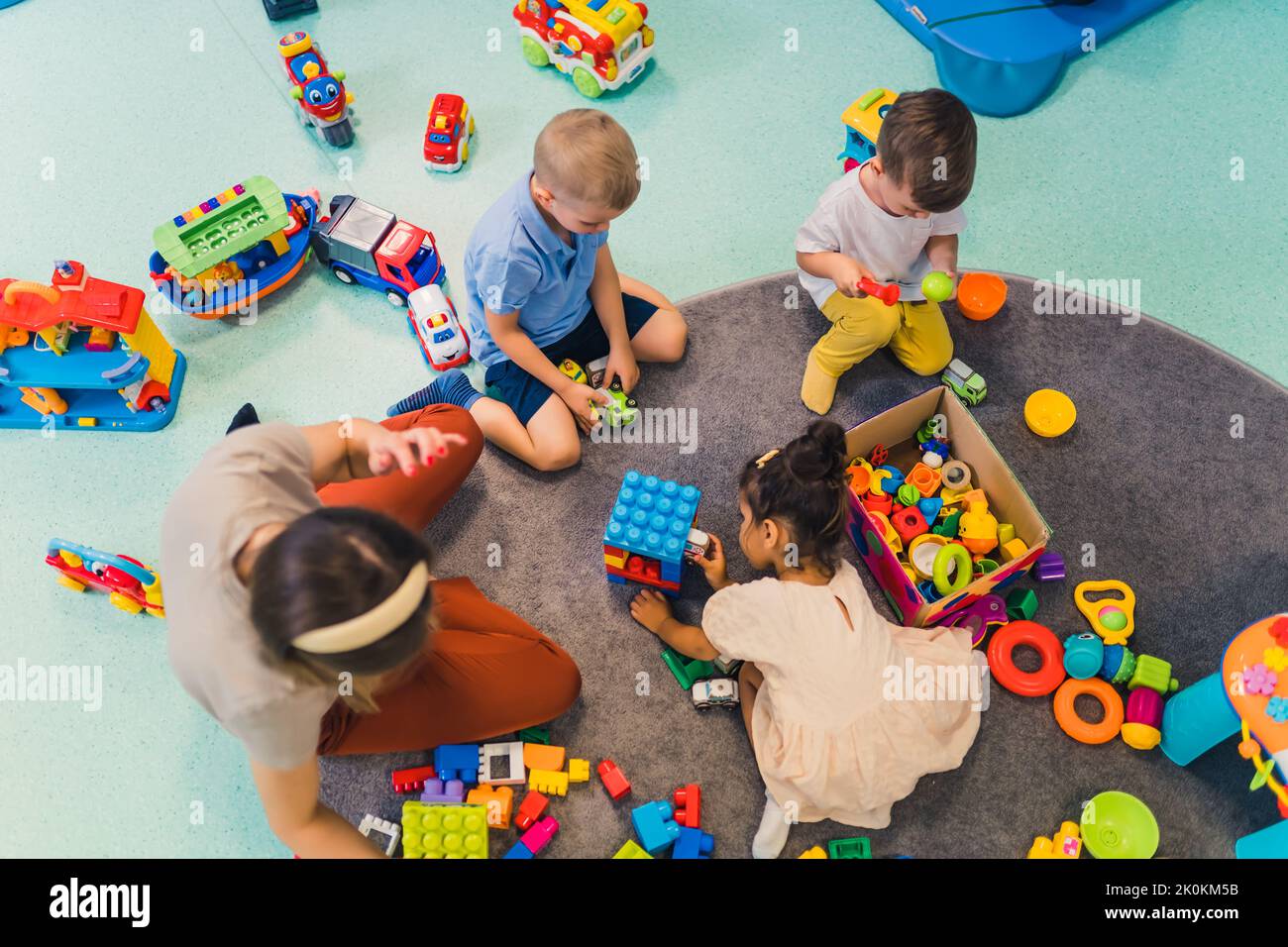 Toddlers and their nursery teacher playing with plastic building blocks and colorful car toys while sitting on the floor in a playroom. Early brain and skills development. High quality photo Stock Photo