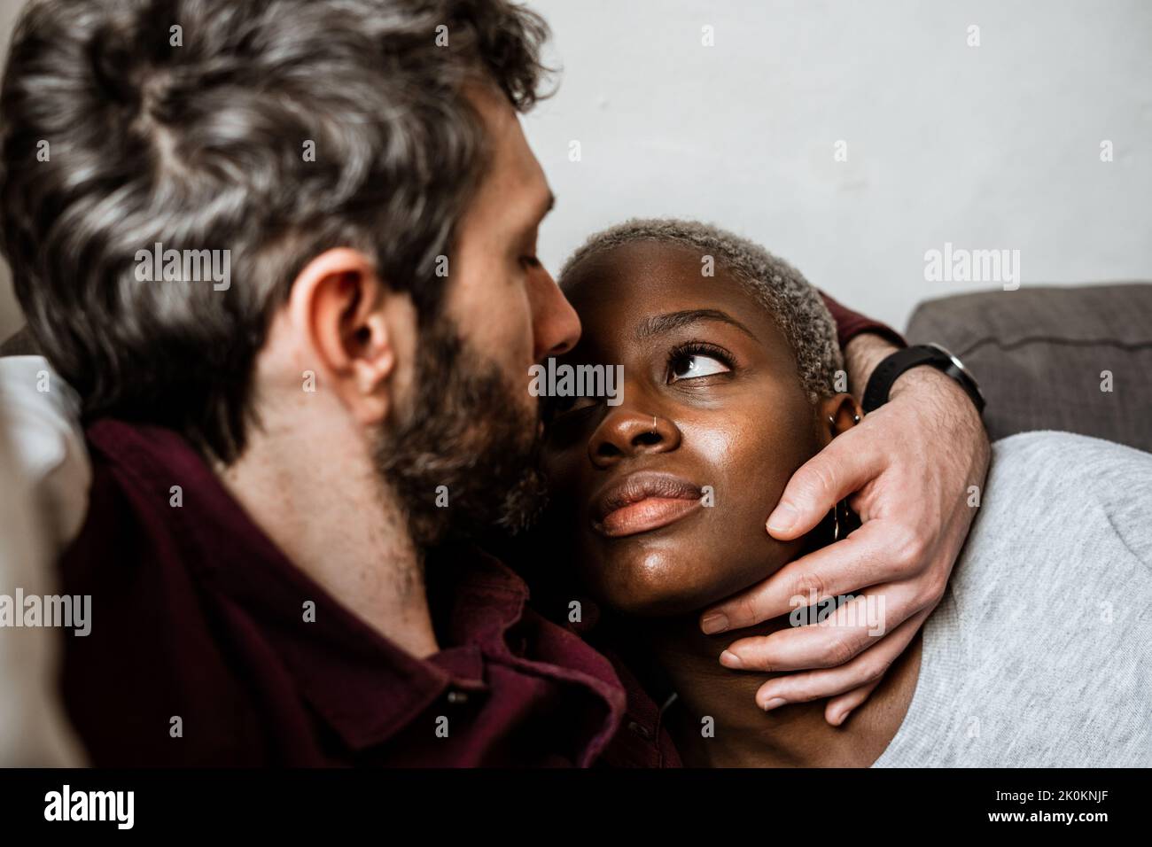 Young bearded man in casual shirt lying on comfortable couch in embrace with African American woman in cozy living room looking at each other Stock Photo