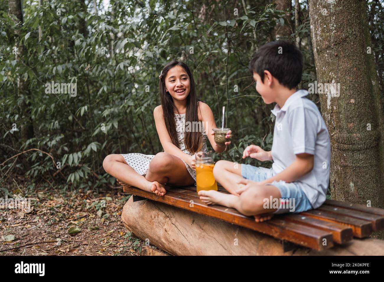 Cheerful barefoot child passing calabash gourd of herbal tea to sibling while looking at each other on bench in summer Stock Photo