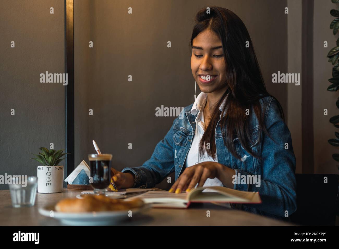 Content smart young Hispanic female student writing in copybook while doing homework assignment at table drinking coffee Stock Photo