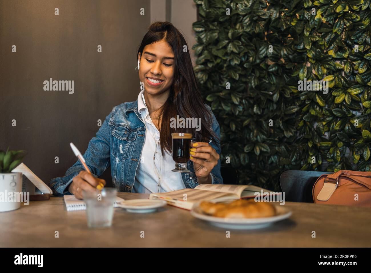 Content smart young Hispanic female student writing in copybook while doing homework assignment at table drinking coffee Stock Photo