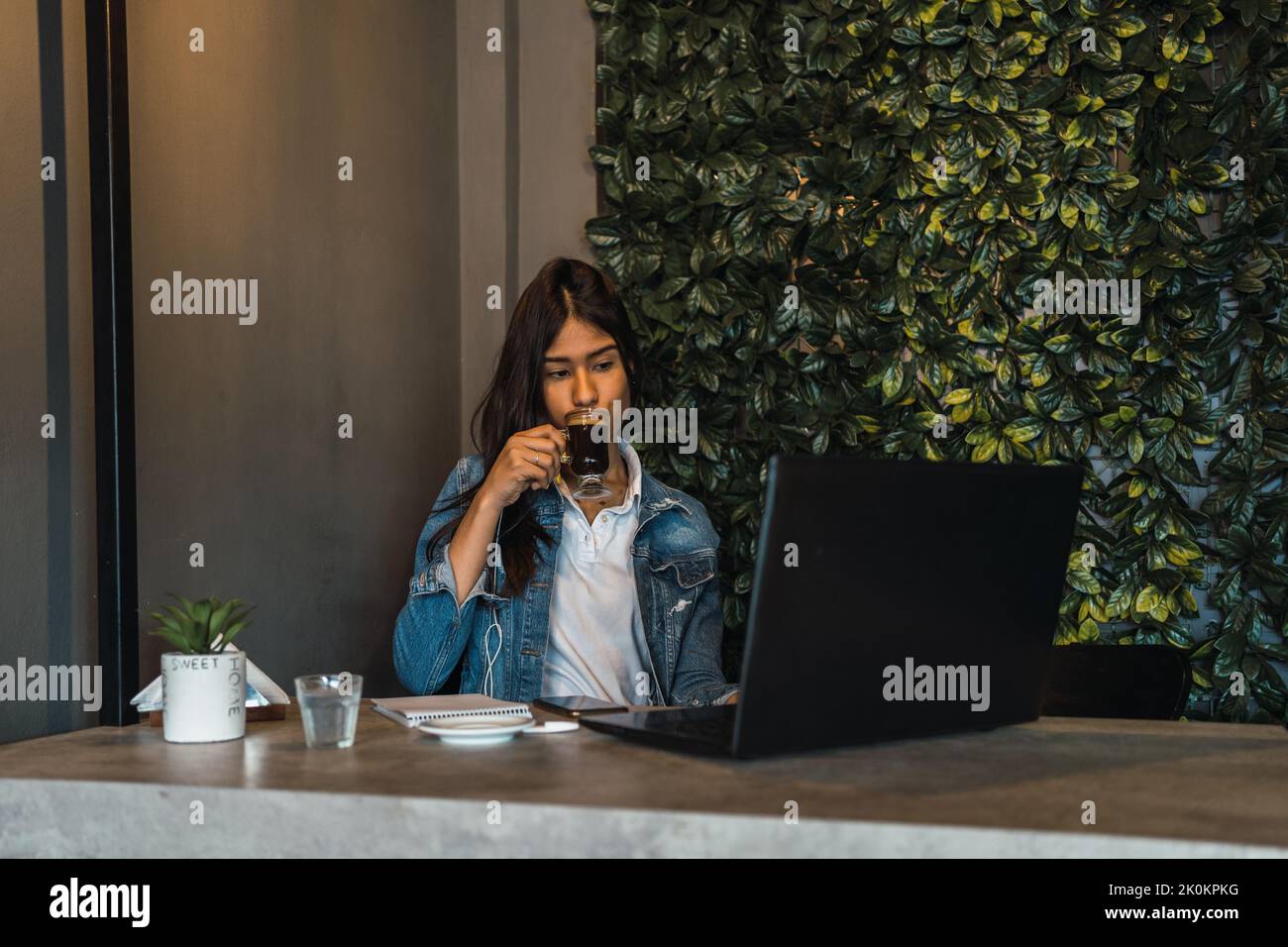 Young Hispanic female freelancer drinking coffee and using laptop while working on remote project at table in cafe Stock Photo