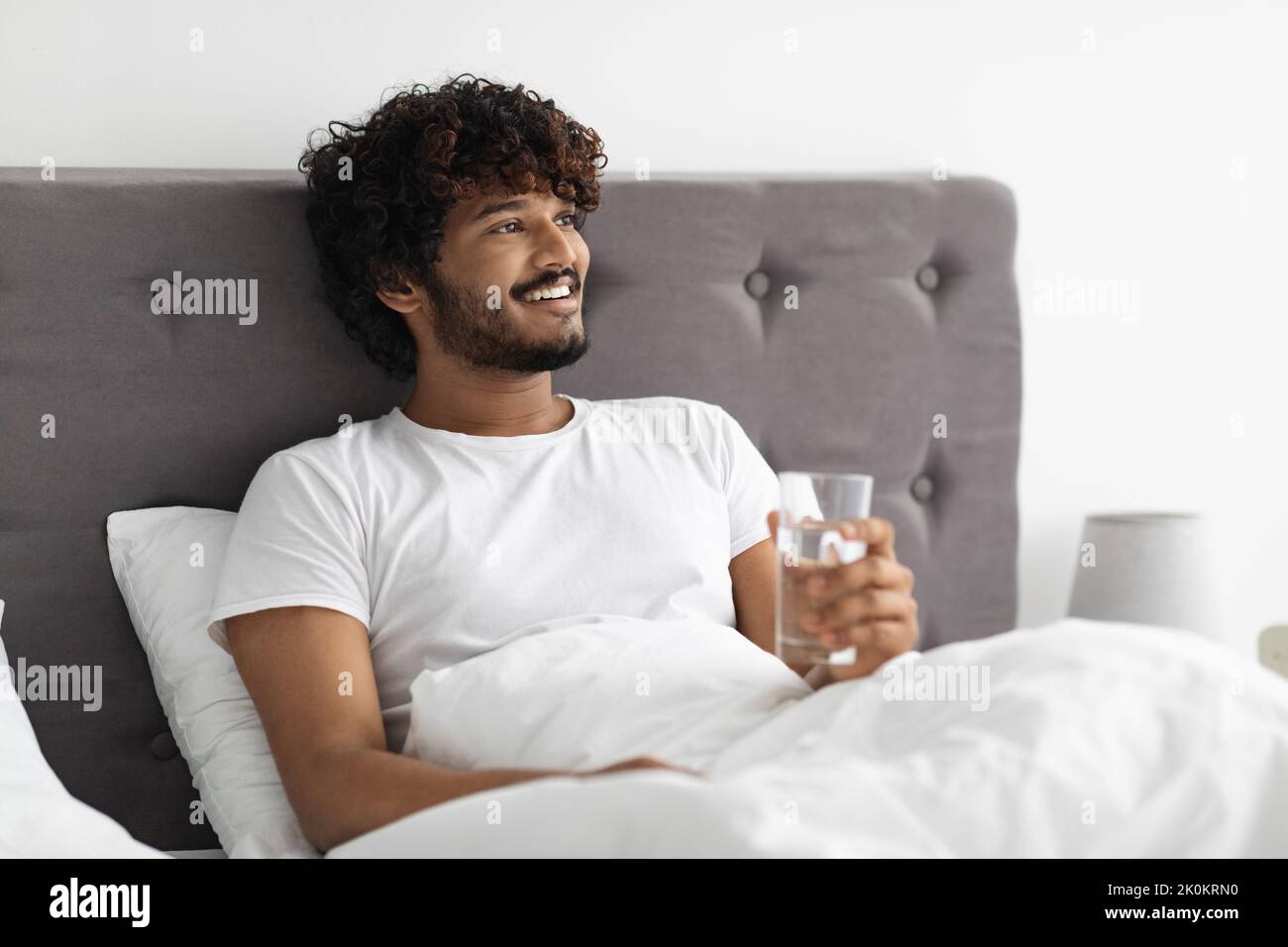 Happy young indian man holding glass of water, bedroom interior Stock ...