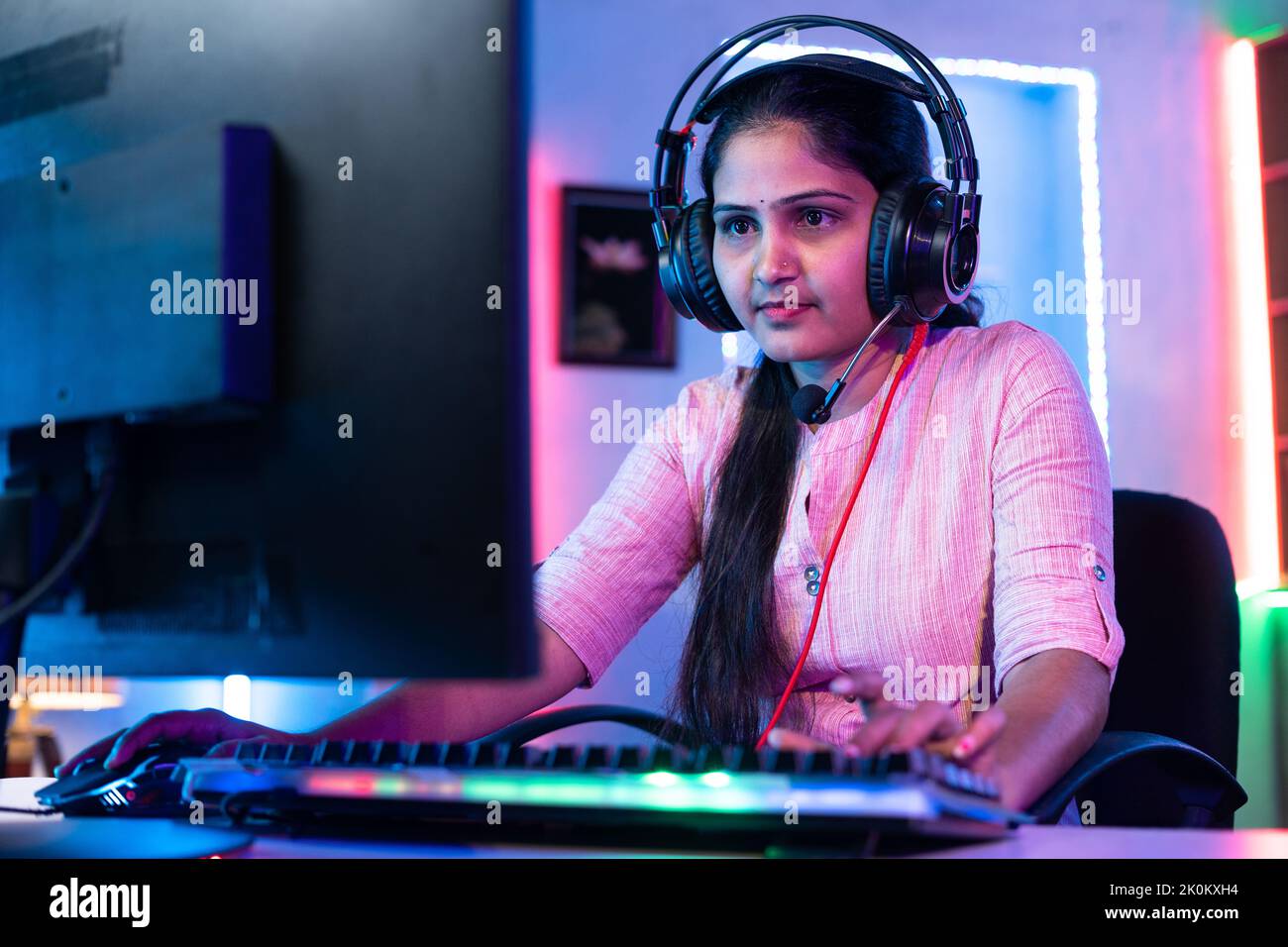 Smiling girl playing online live video game on computer by talking on headphone at home - concept of social media sharing, championship and gaming Stock Photo