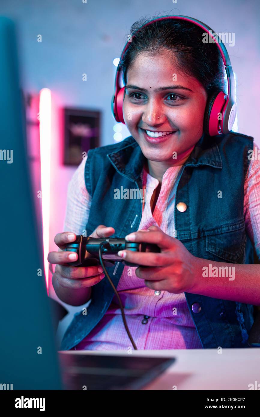 Vertical shot, Smiling girl playing live video game on laptop by talking on headphones at home on neon background - concept of vlogger, gaming Stock Photo