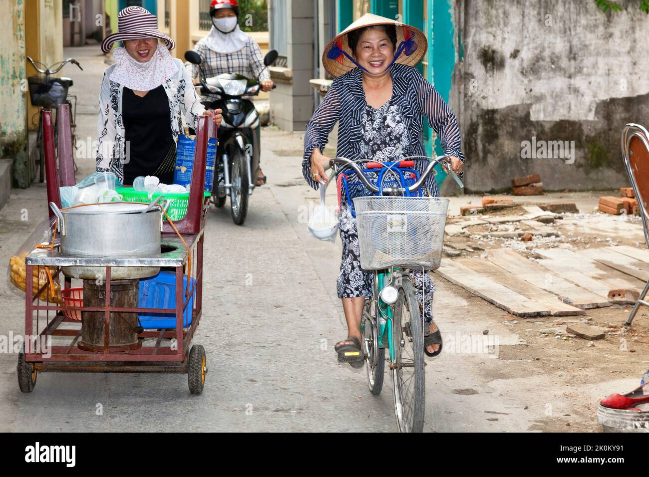 Vietnamese lady wearing bamboo hat on bicycle in narrow alley, Hai Phong, Vietnam Stock Photo