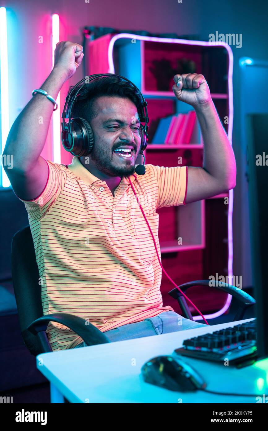 vertical shot of Cheerful gamer celebrating win by raising hands while playing online video game at home - concept of achievement, champion and Stock Photo