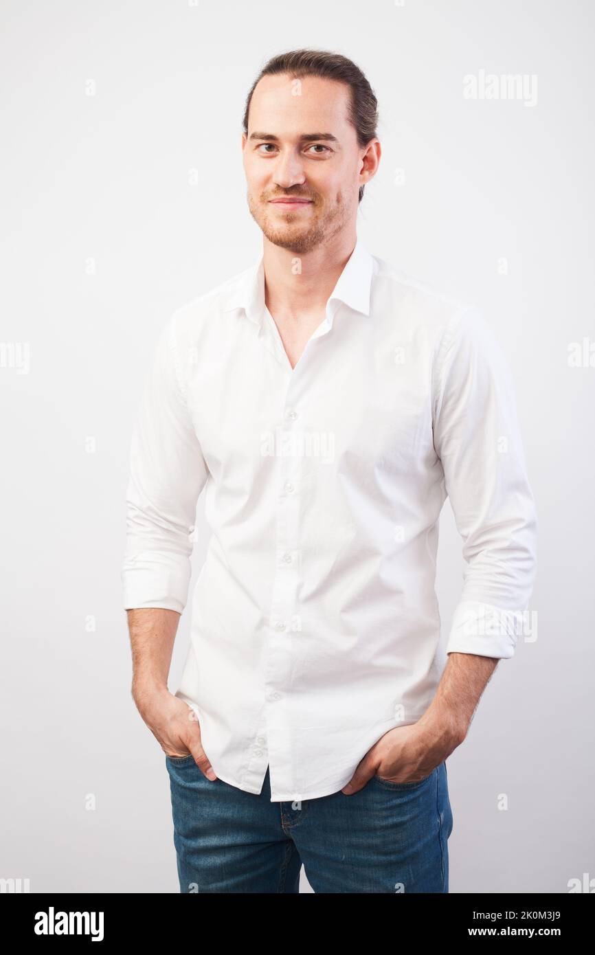 Portrait of young successful young man in white shirt, smiling holding hands in pockets over gray wall. Confidence and business concept. Stock Photo