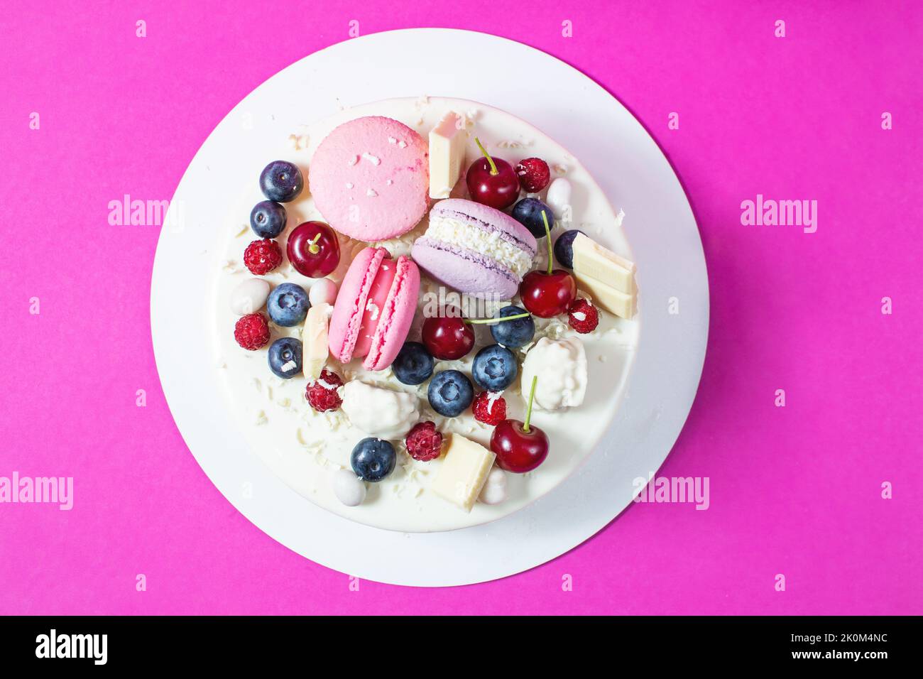 Tender white cake decorated with melted white chocolate, macaroons, meringues, berries and candies on pink background. Top view Stock Photo