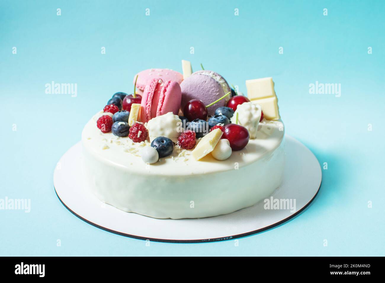 Tender white cake decorated with melted white chocolate, macaroons, meringues, berries and candies on blue background. Stock Photo