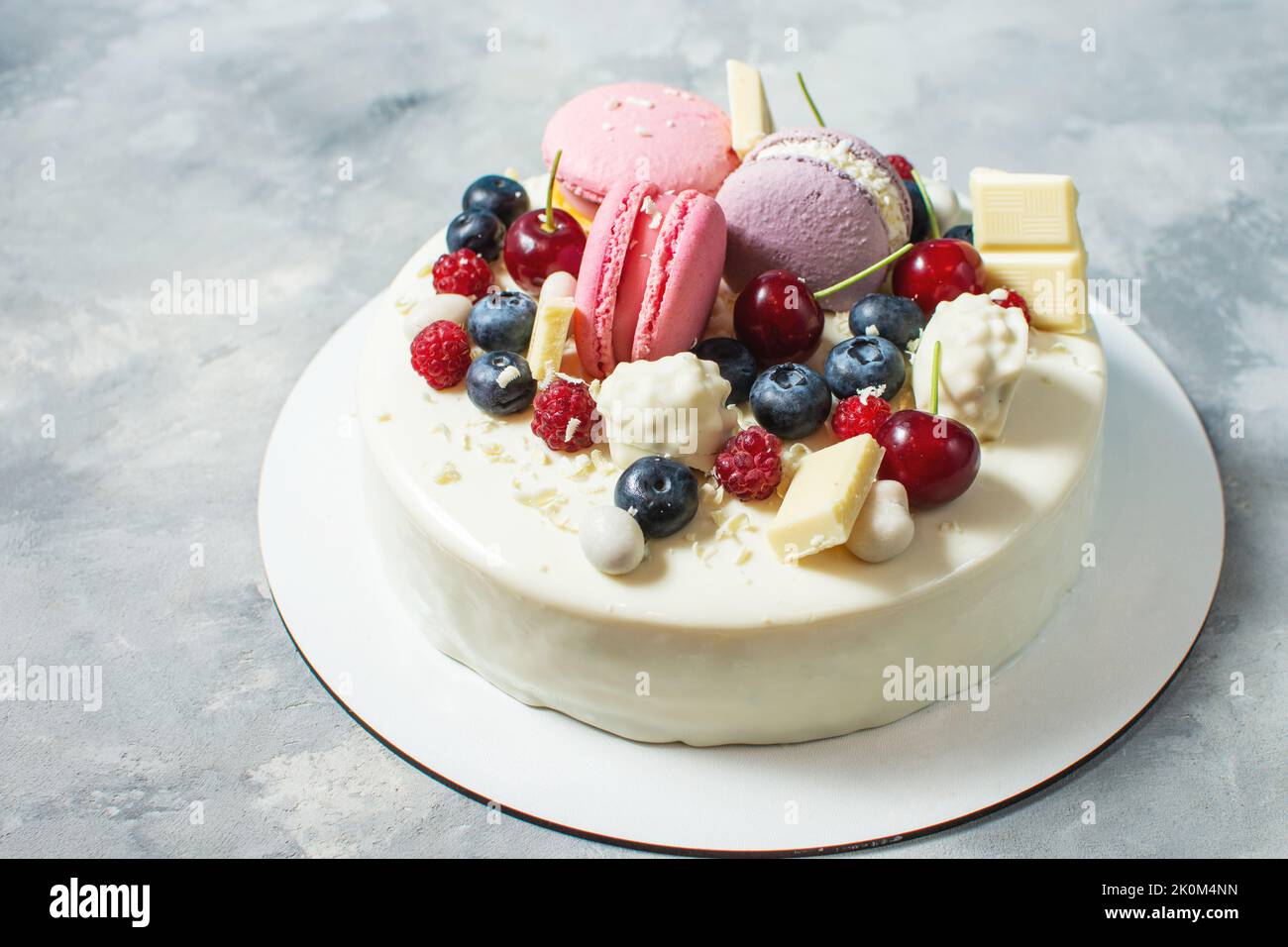 Tender white cake decorated with melted white chocolate, macaroons, meringues, berries and candies on concrete background. Stock Photo