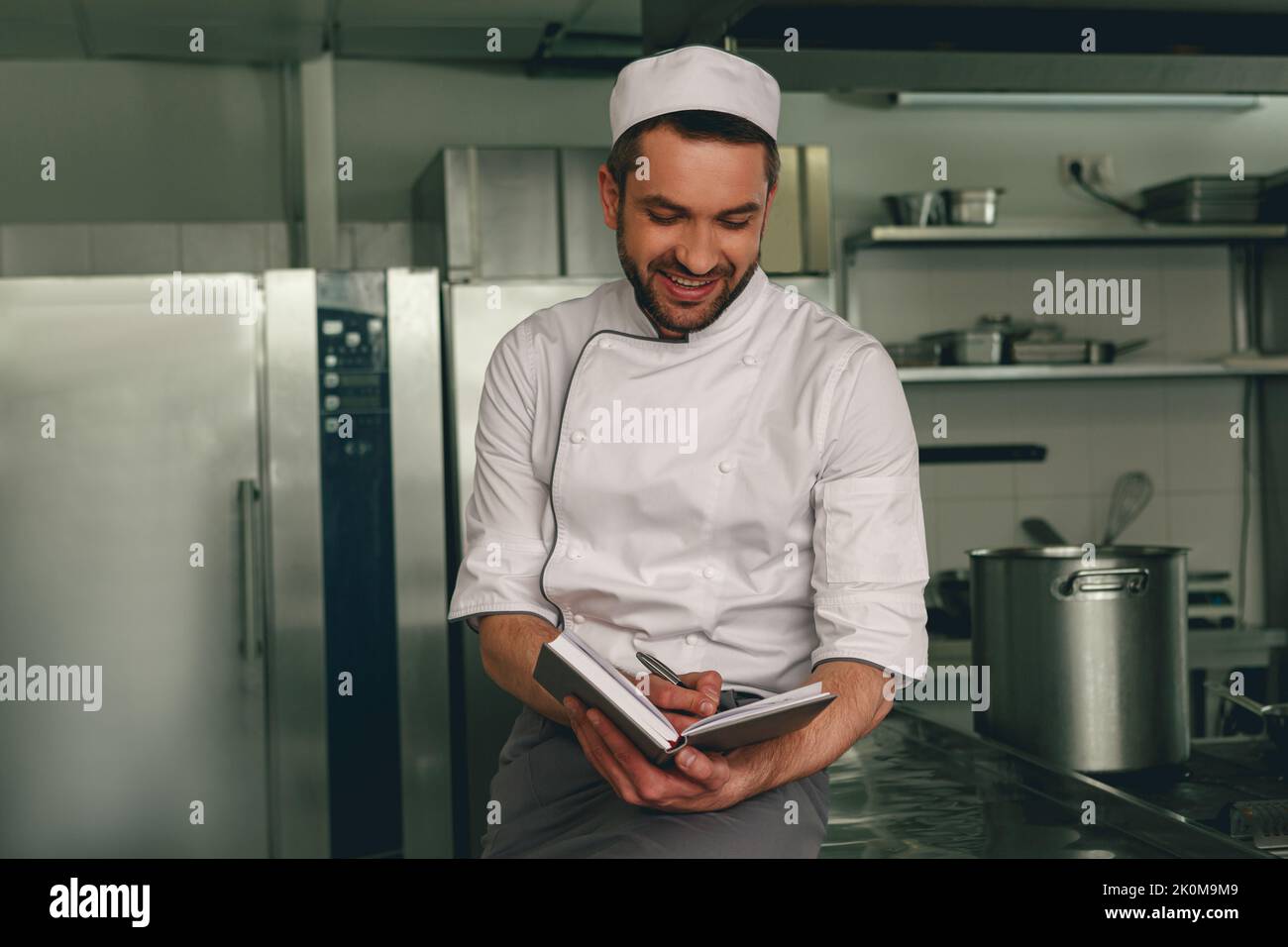 Smiling chef in uniform making notes in notebook standing on kitchen Stock Photo