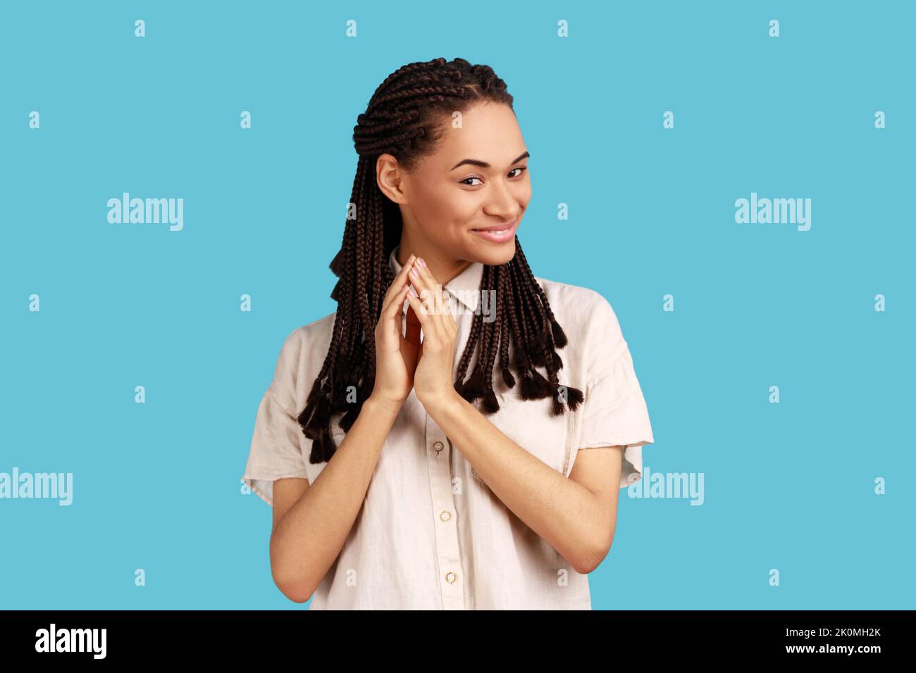 Portrait of thoughtful woman with black dreadlocks schemes something, keeps fingers together, considers over cunning plans, wearing white shirt. Indoor studio shot isolated on blue background. Stock Photo