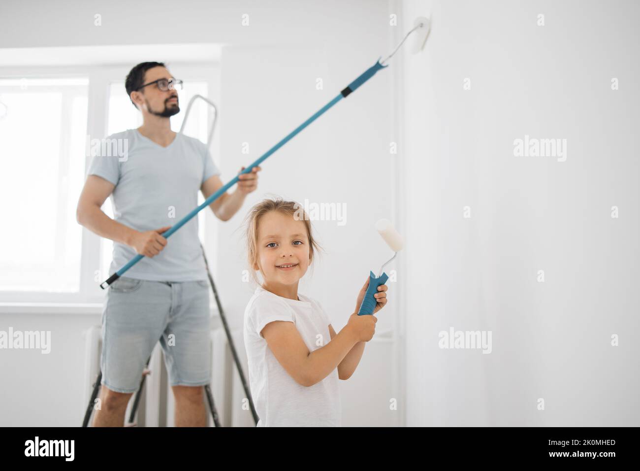 Smiling 4-year-old girl holds a paint roller in her hands. The child helps her father, who is standing behind her, near the wall, in the repair. Stock Photo