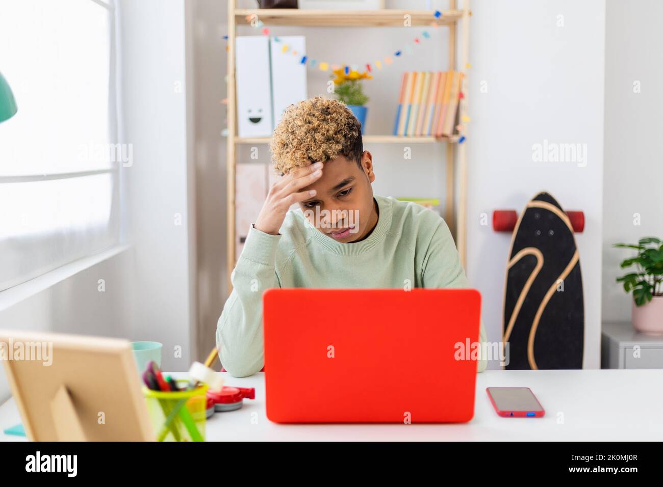 Stressed teenager boy sitting at desk studying on laptop in room Stock Photo
