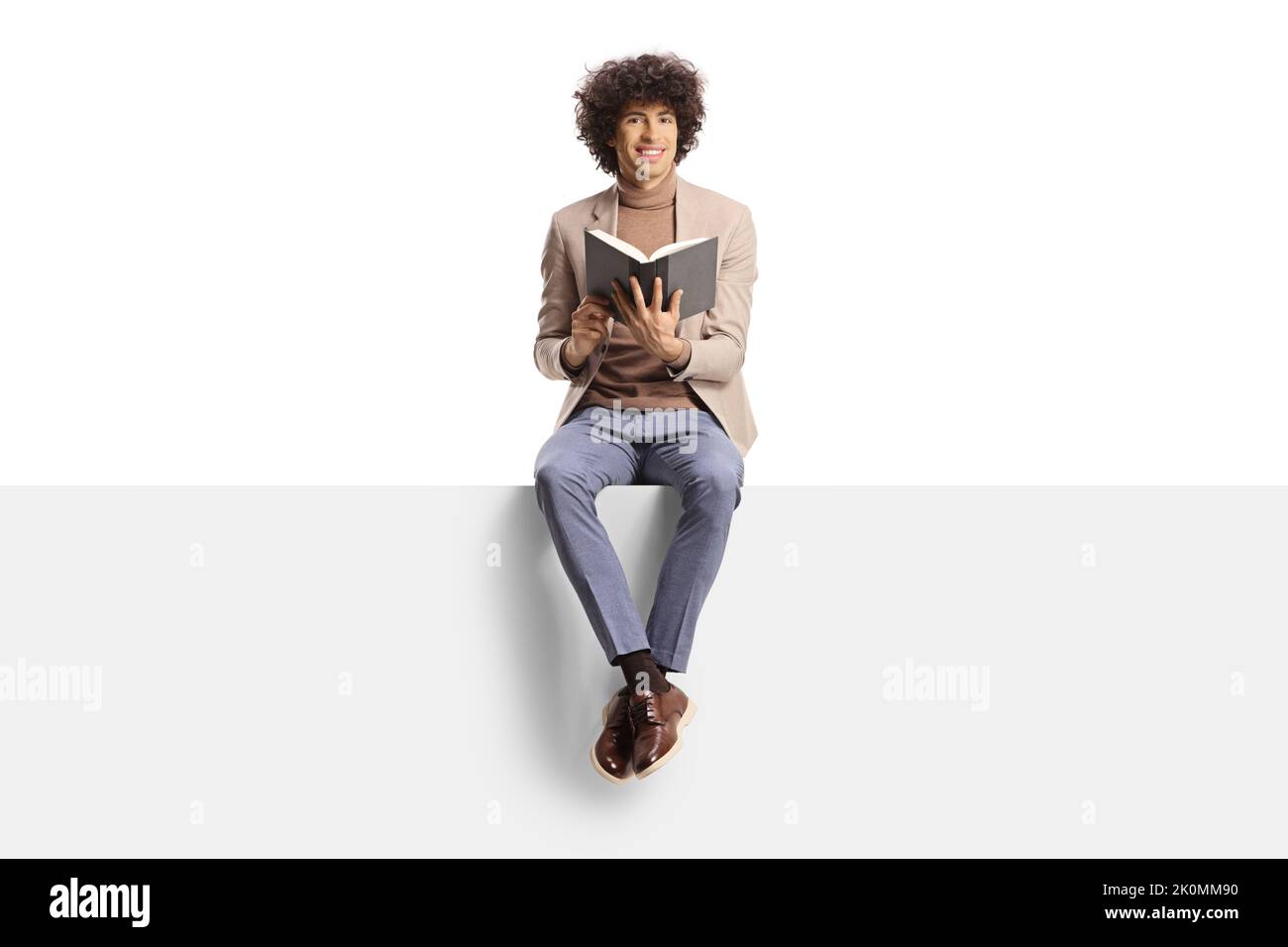 Elegant young man with curly hair sitting on a panel and holding a book isolated on white background Stock Photo