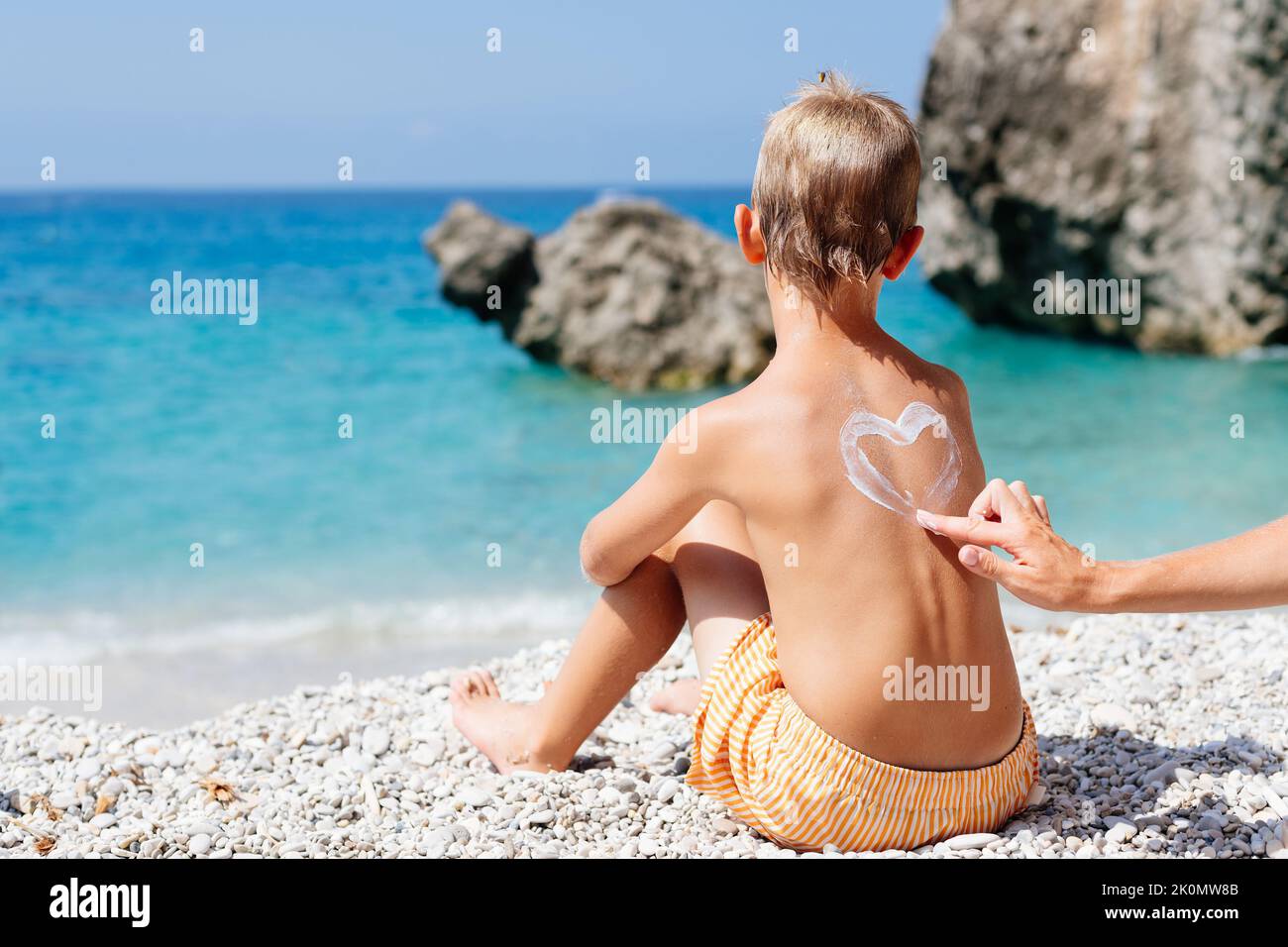 Mom smears her baby's back with sunscreen on a beach in Corfu, Greece. She draws a heart of cream on her son's back.  Stock Photo