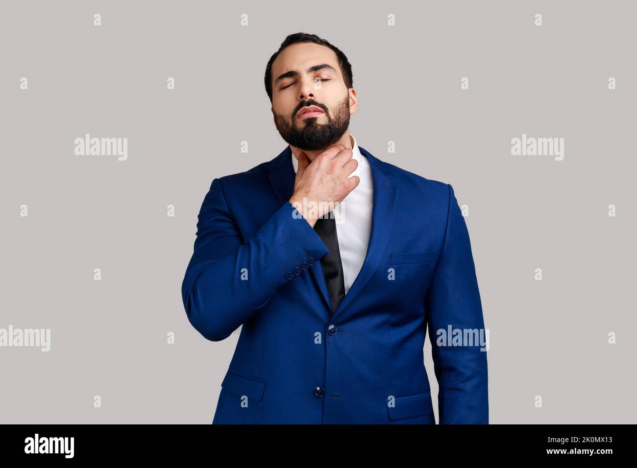 Unhealthy bearded man touching neck feeling pain while swallowing, result of chocking, throat inflammation, sore throat, wearing official style suit. Indoor studio shot isolated on gray background. Stock Photo