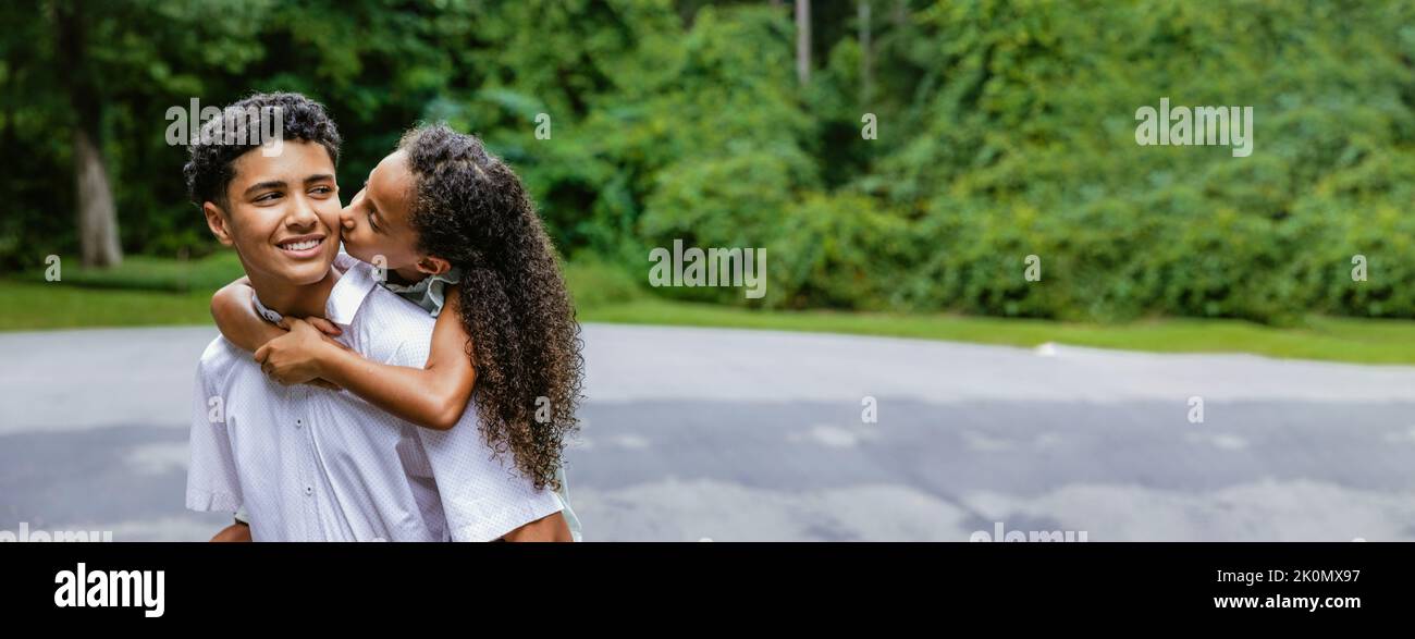 A little sister on her big teenage brother's back giving him a kiss on the cheek with copy space. Stock Photo