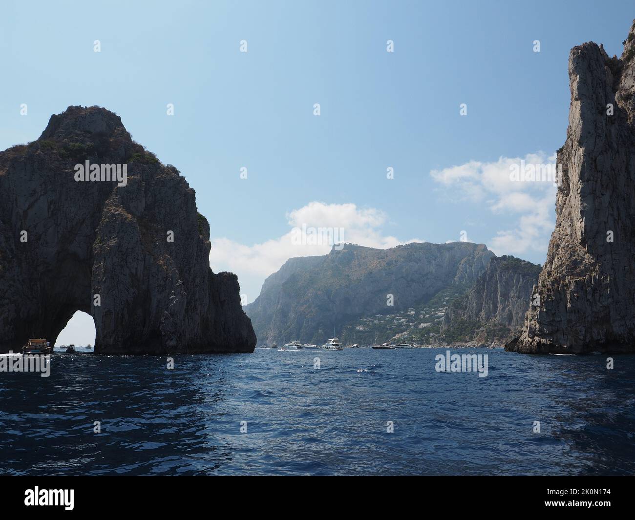 The coast of Capri island is very rocky and steep. These are the iconic Faraglioni rocks, symbols of the island. Tourist boats pass through the arch. Stock Photo
