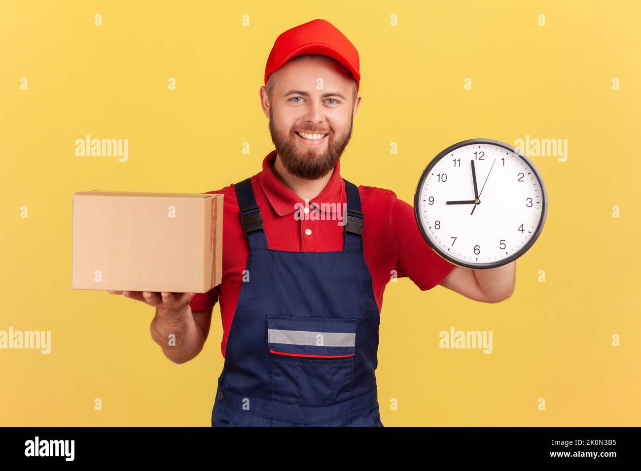 Food delivery, shipping on time. Courier man wearing overalls and T-shirt holding clock and cardboard box, express courier transportation. Indoor studio shot isolated on yellow background. Stock Photo