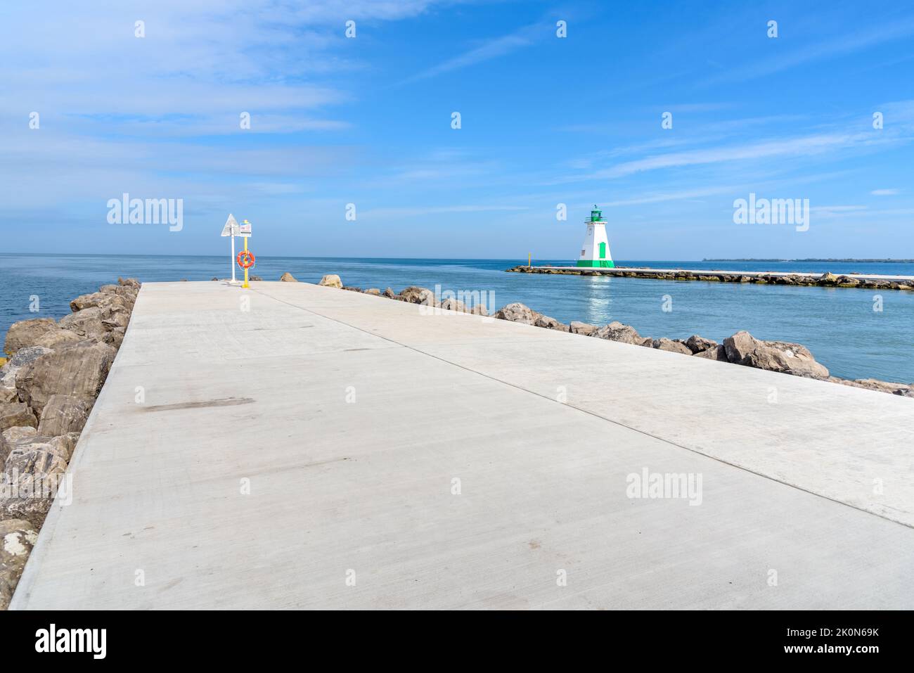 Concrete path on the breakwater at the entrance of a harbour on a lake. A lighthouse is on the breakwater at the other side of the canal. Lake Ontario Stock Photo
