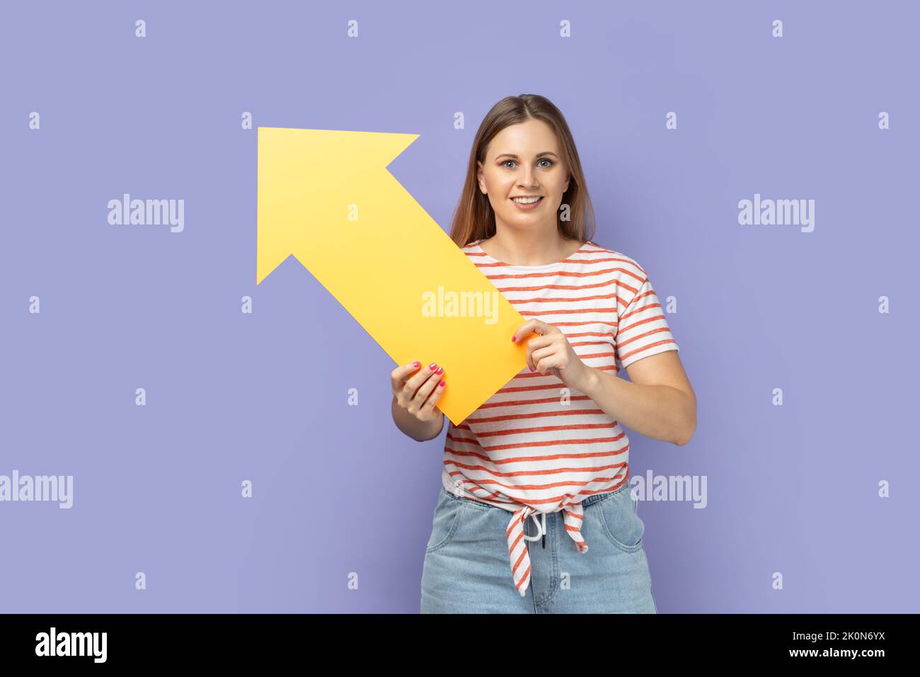 Portrait of delighted cute blond woman wearing striped T-shirt holding big yellow arrow indicating aside, looking at camera with smile. Indoor studio shot isolated on purple background. Stock Photo