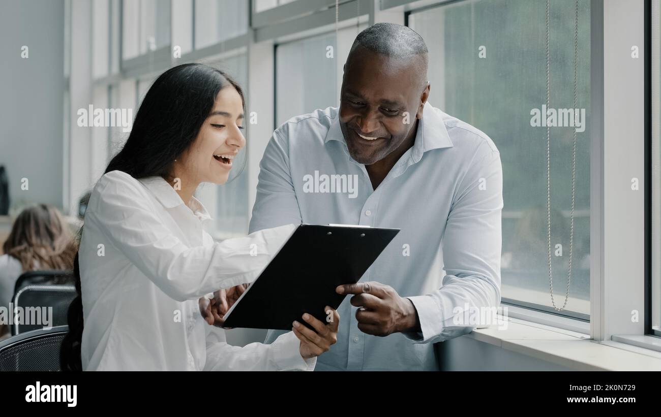 Diverse african and arabian colleagues multiracial people business partners stand near window discussing paper graphics strategic plans reading Stock Photo