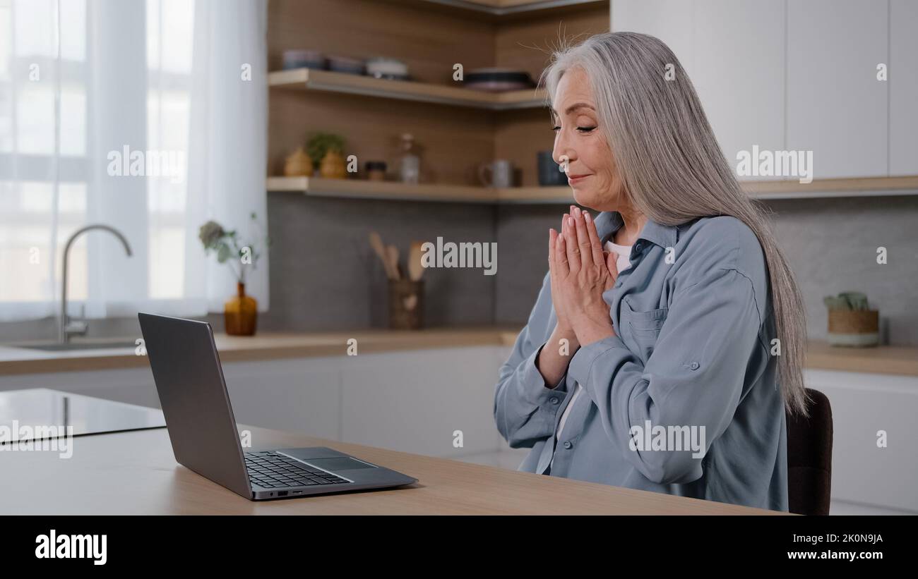 Caucasian senior woman elderly grey-haired lady sit in kitchen talk at computer webcam wave hand hello gesture video call conference chat with family Stock Photo