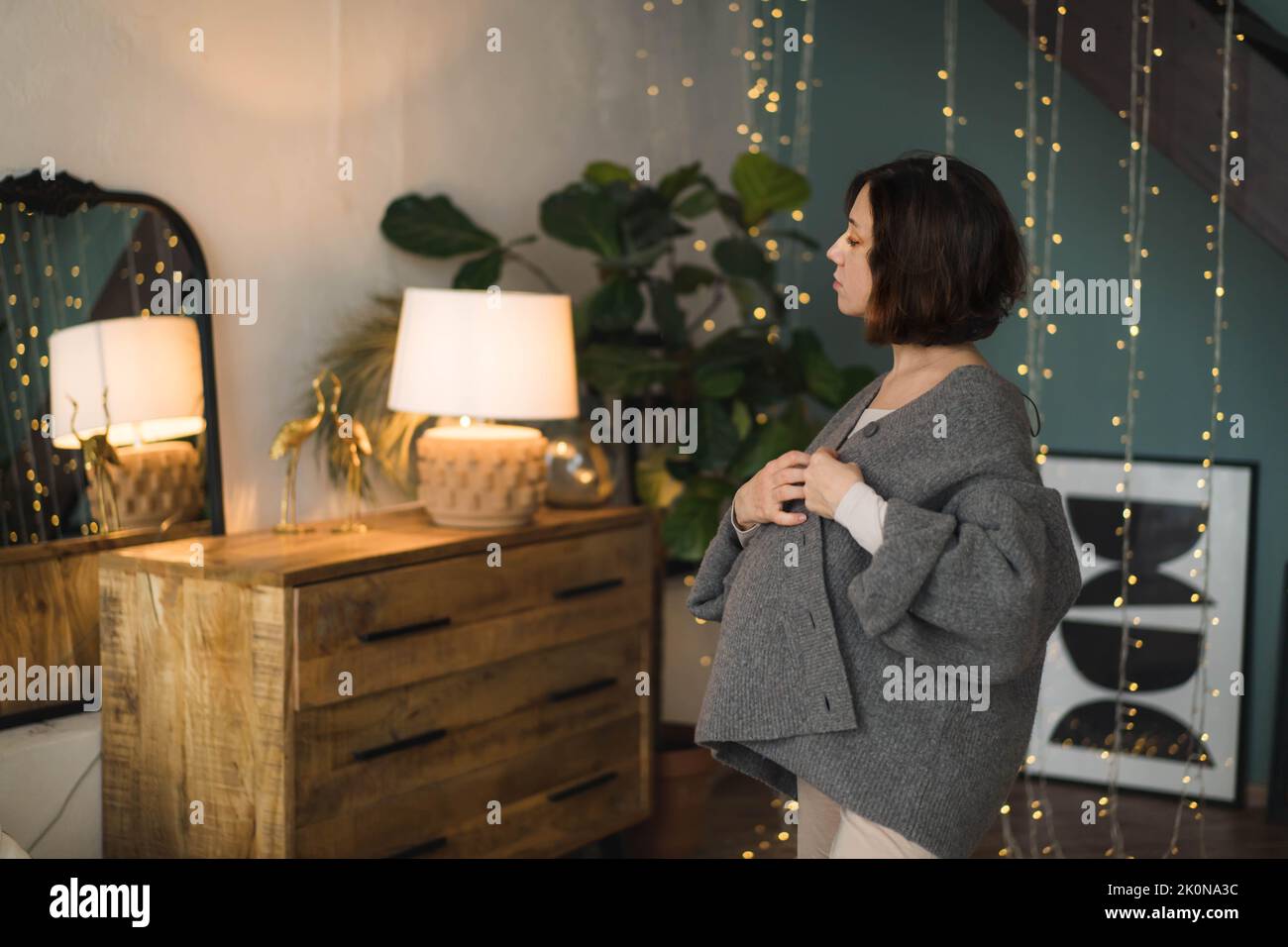 A young pregnant woman looks at herself in the mirror. Close-up portrait photo of a pregnant woman in a warm knitted sweater . Concept of happy pregnancy, motherhood, expecting baby. Stock Photo