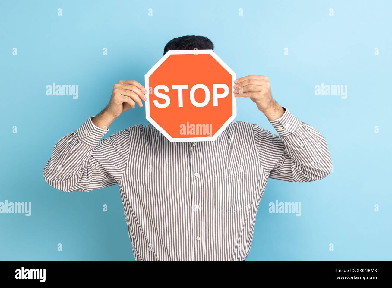 Portrait of man covering face with Stop symbol, anonymous person holding red traffic sign, warning to go, prohibition concept, wearing striped shirt. Indoor studio shot isolated on blue background. Stock Photo