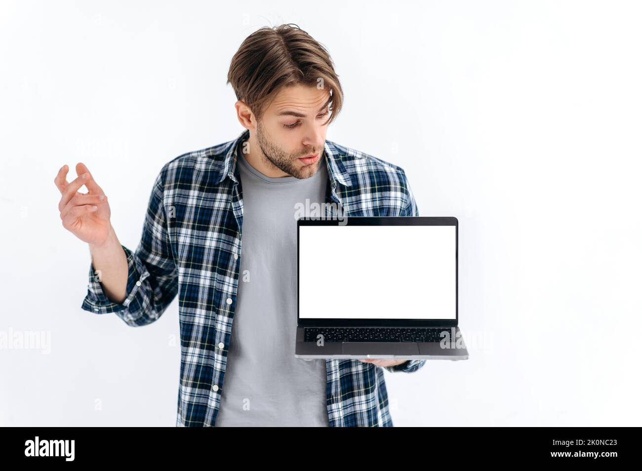Mock-up concept. Confused puzzled caucasian guy in casual wear, freelancer or student, holding an open laptop with empty mock-up space, peeks at it in surprised, stand on white isolated background Stock Photo
