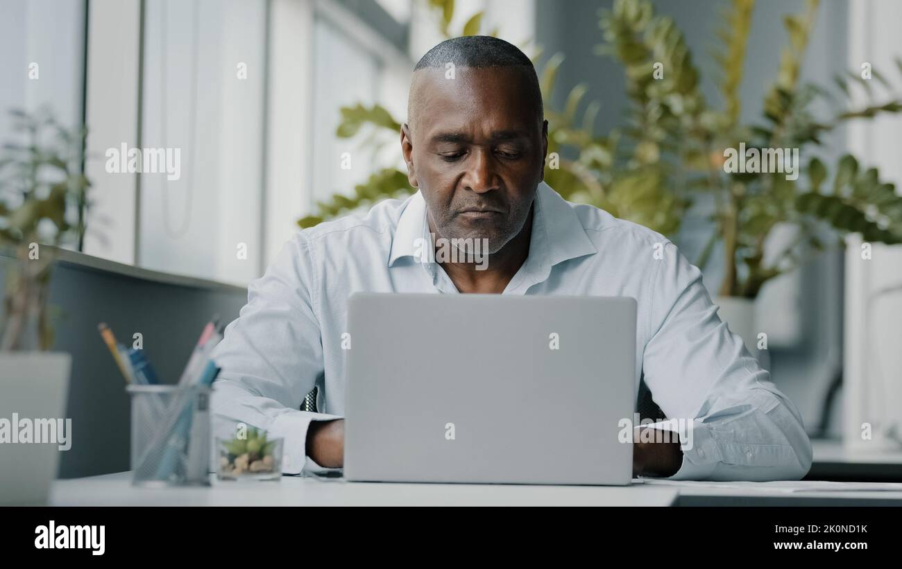 Concentrated african man employee american expert focused specialist sits in office works on laptop use personal computer typing email chatting Stock Photo