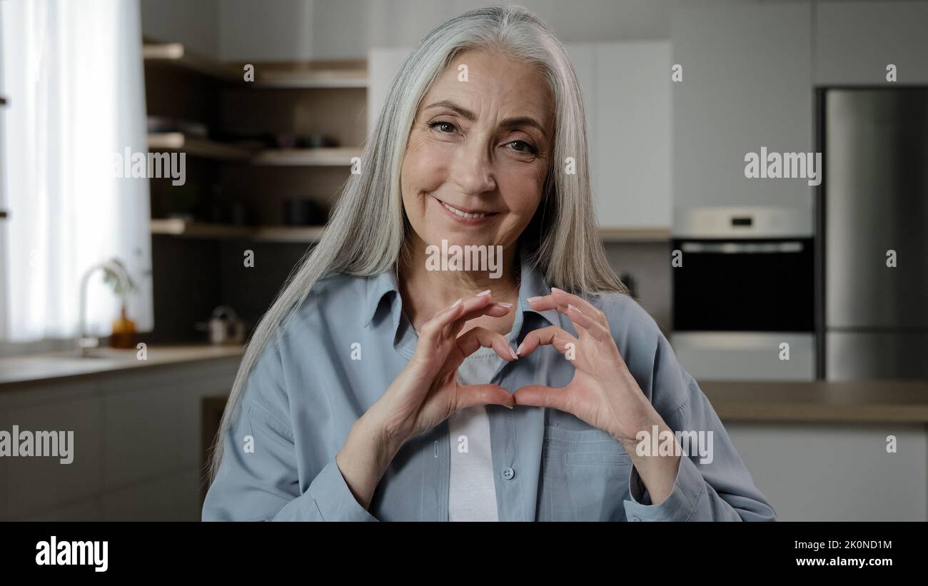 Kindly thankful 60s woman elderly mature volunteer female pensioner gray-haired granny sweetheart makes romantic love frame by fingers heart form Stock Photo
