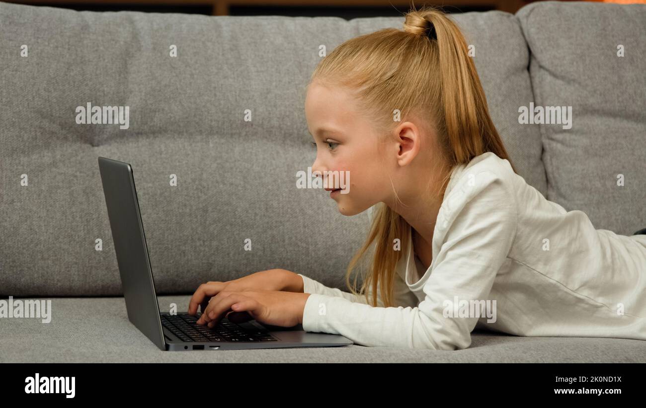 Carefree little kid pretty blonde girl child daughter using laptop lying on grey sofa surfing internet chatting with classmates playing games studying Stock Photo