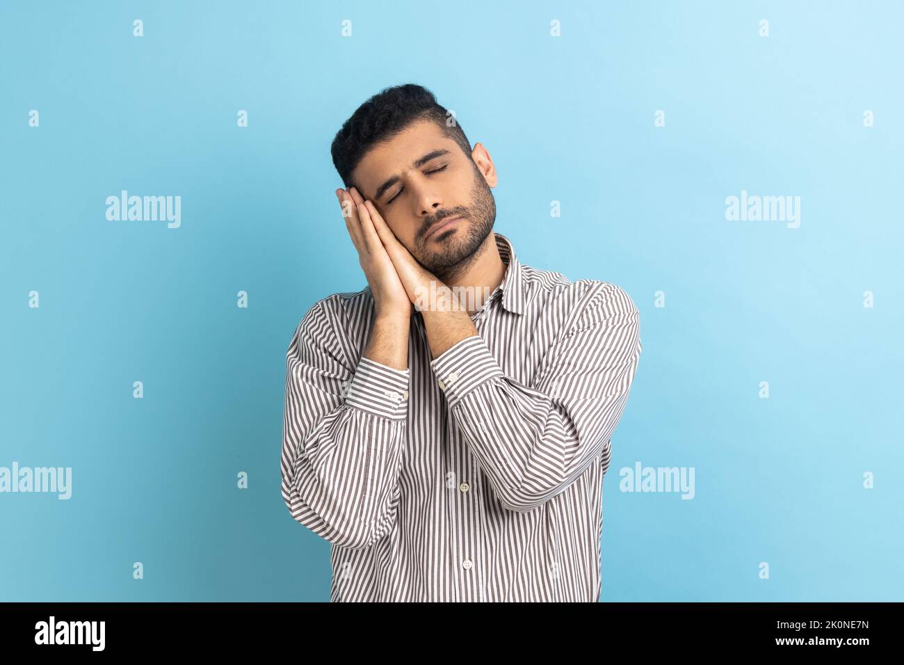 Portrait of handsome bearded man sleeping laying down on her palms, having comfortable nap and resting, dozing off, wearing striped shirt. Indoor studio shot isolated on blue background. Stock Photo