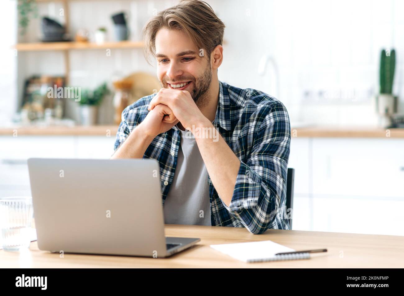 Positive successful modern caucasian male freelancer, programmer, stylishly dressed, sits at desk with laptop in a kitchen at home, working on a creative project, looking to the side, smiling happily Stock Photo
