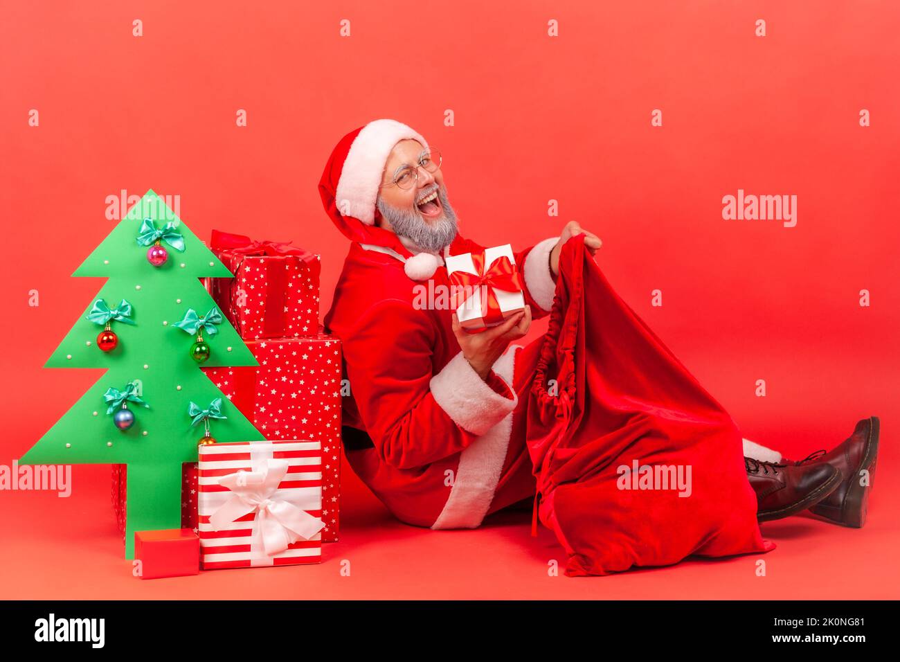 Happy positive elderly man with gray beard wearing santa claus costume with festive mood, taking present box from bag, looking at camera and smiling. Indoor studio shot isolated on red background. Stock Photo